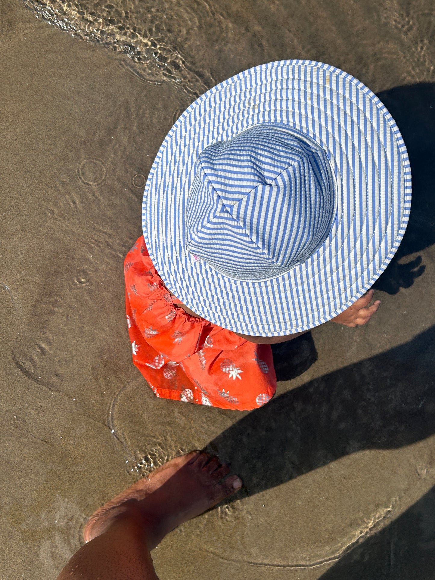 Toddler wearing Blue and white striped sun hat on beach with coral beach dress with pineapple motifs